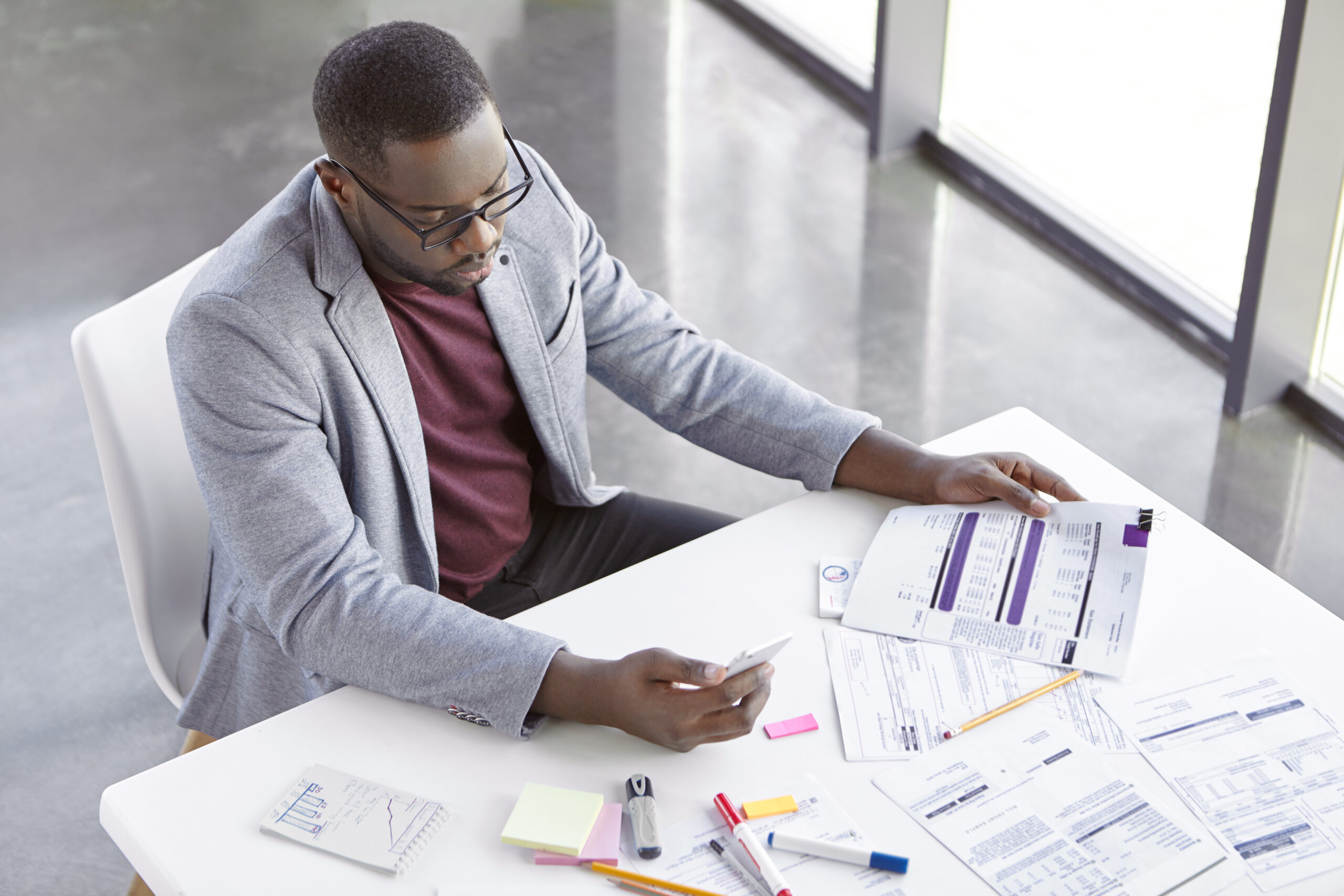 A stressed Black retail business owner holding paper invoices and a smartphone while manually managing company finances at a white office desk.