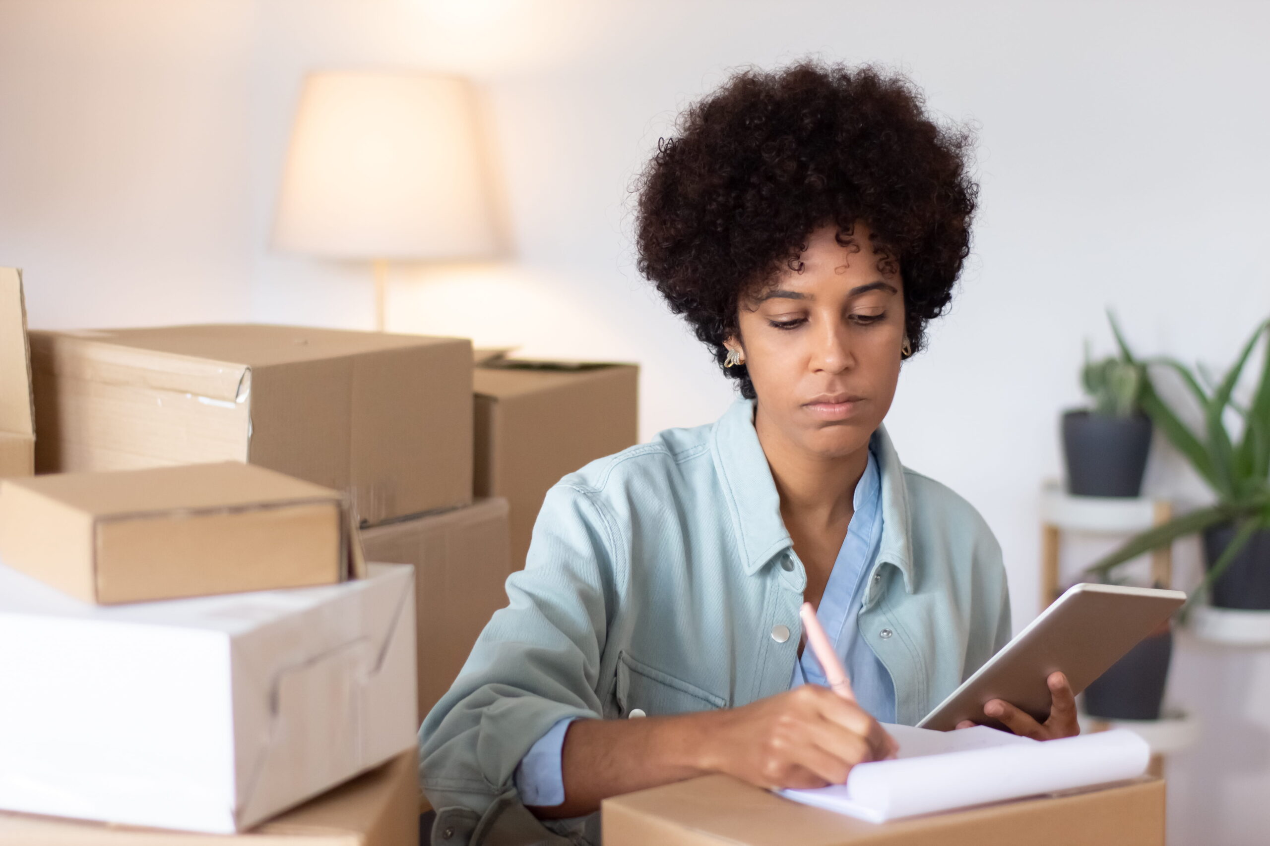 Female retailer managing stock control by hand with notebook and tablet in a storeroom